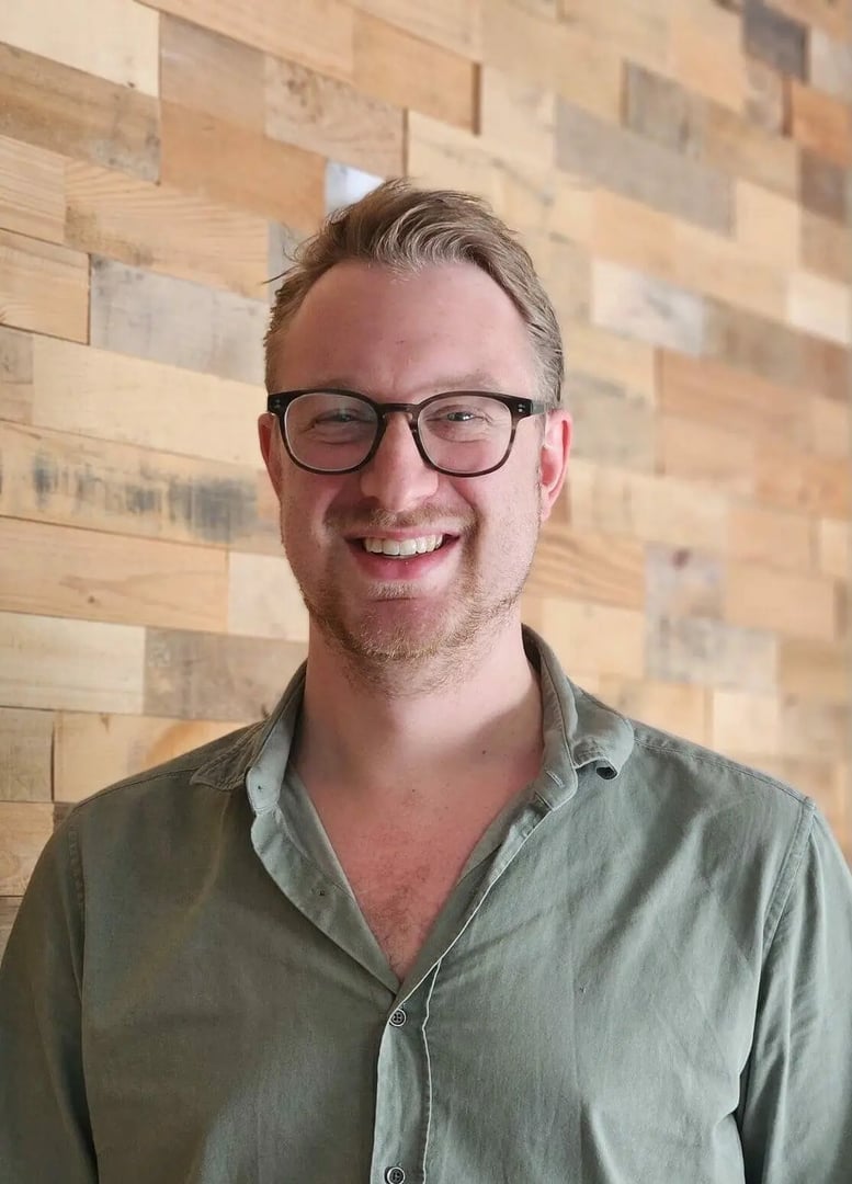 Smiling person wearing glasses and a green shirt stands in front of a wooden wall.