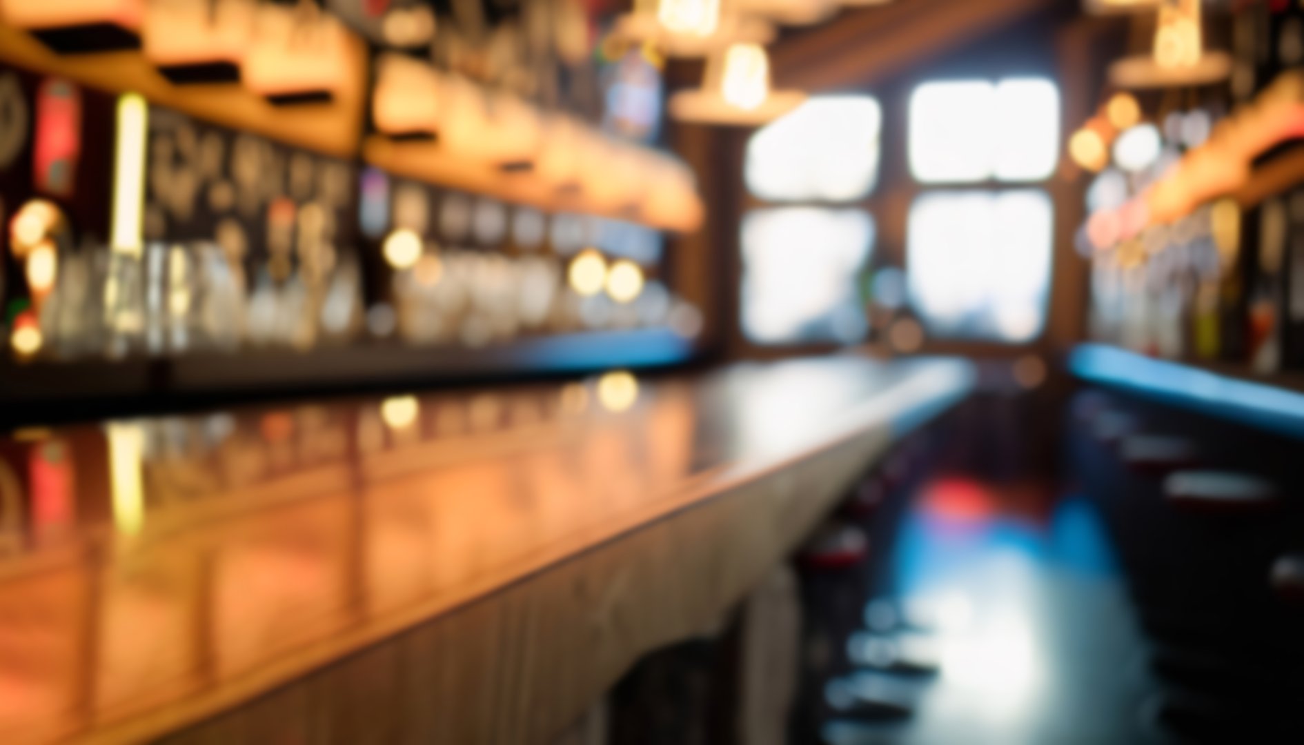 Blurred image of an empty, stylish bar with warm ambient lighting. The focus is on the wooden bar counter, with hanging lights and bar stools slightly visible in the background, creating a cozy atmosphere.