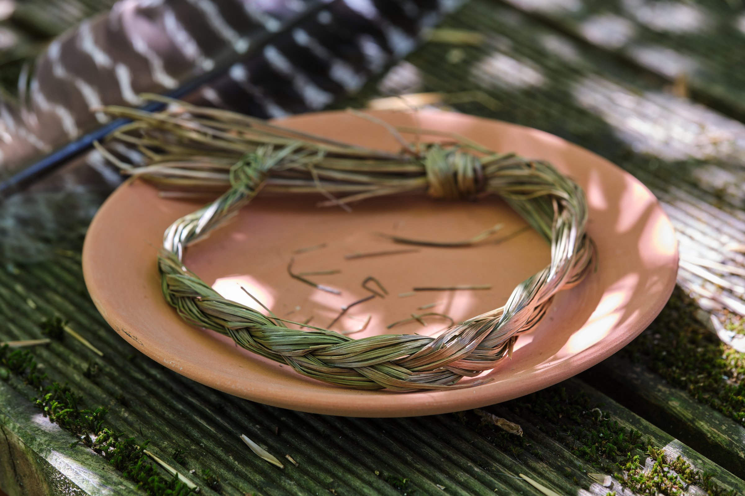 Sweetgrass braid (Hierochloe odorata), also called vanilla grass, holy grass or mary's grass, and smudging feather on a clay plate. Sweetgrass is widely used by Indigenous peoples from in North America to attract good spirits. Close-up, eye level view, photographed outdoors in summer on a sunny day.