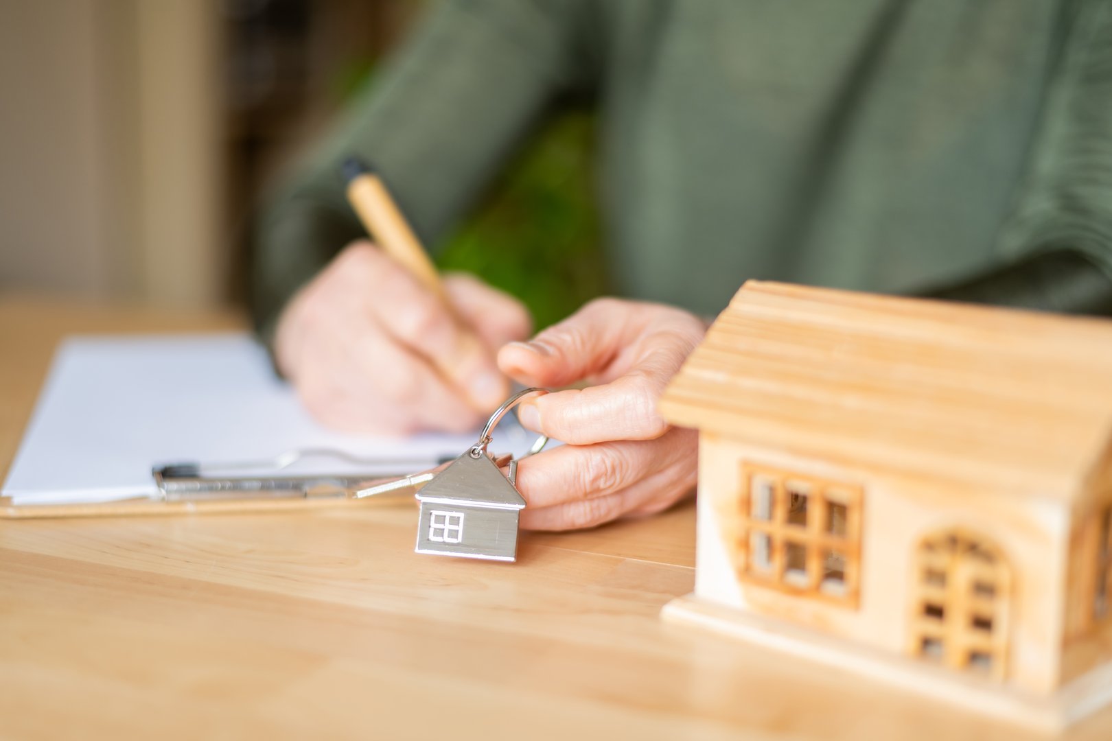 Real estate agent is signing a contract for the sale of a property and holding keys of a new house, with a small wooden house model on the table
