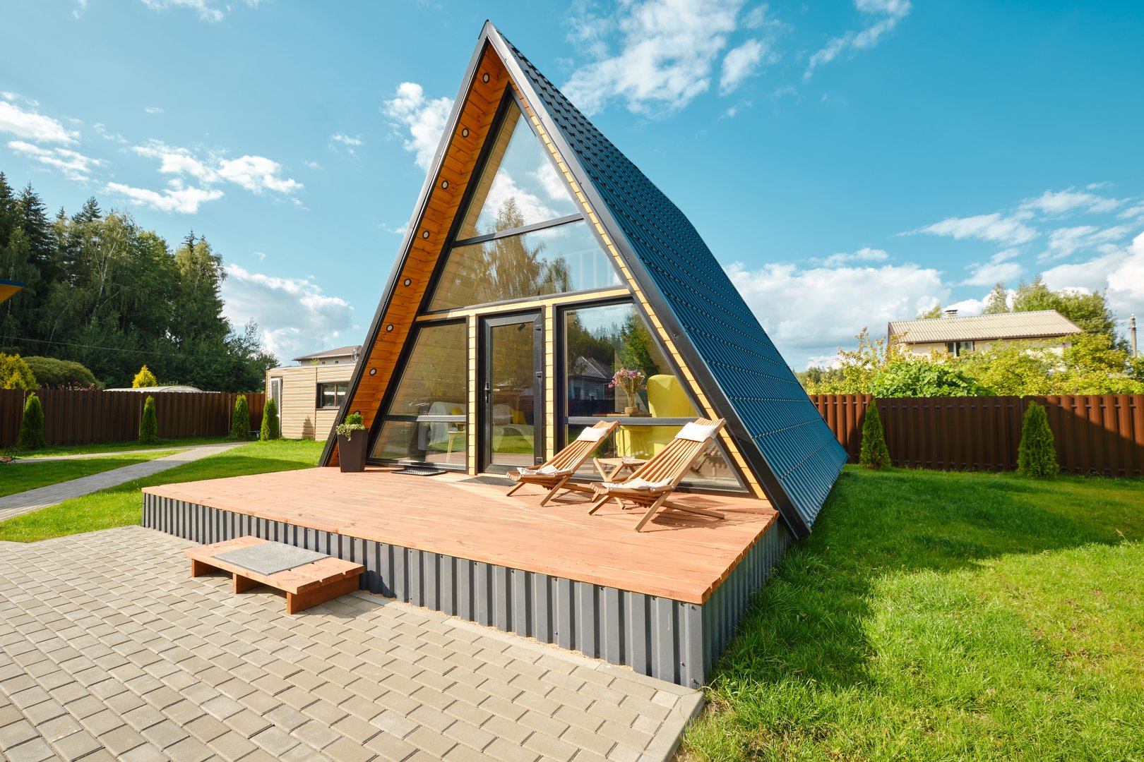 This unique A-frame house features large glass windows and a wooden deck, surrounded by green grass and trees under a bright blue sky. Two lounge chairs sit invitingly on the deck.