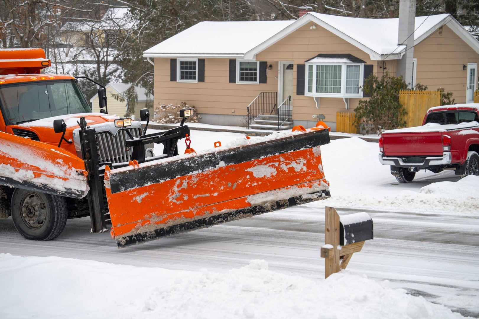 Snow plow truck clearing residential driveway in Anchorage, Alaska, with snow-covered house and red pickup truck in background