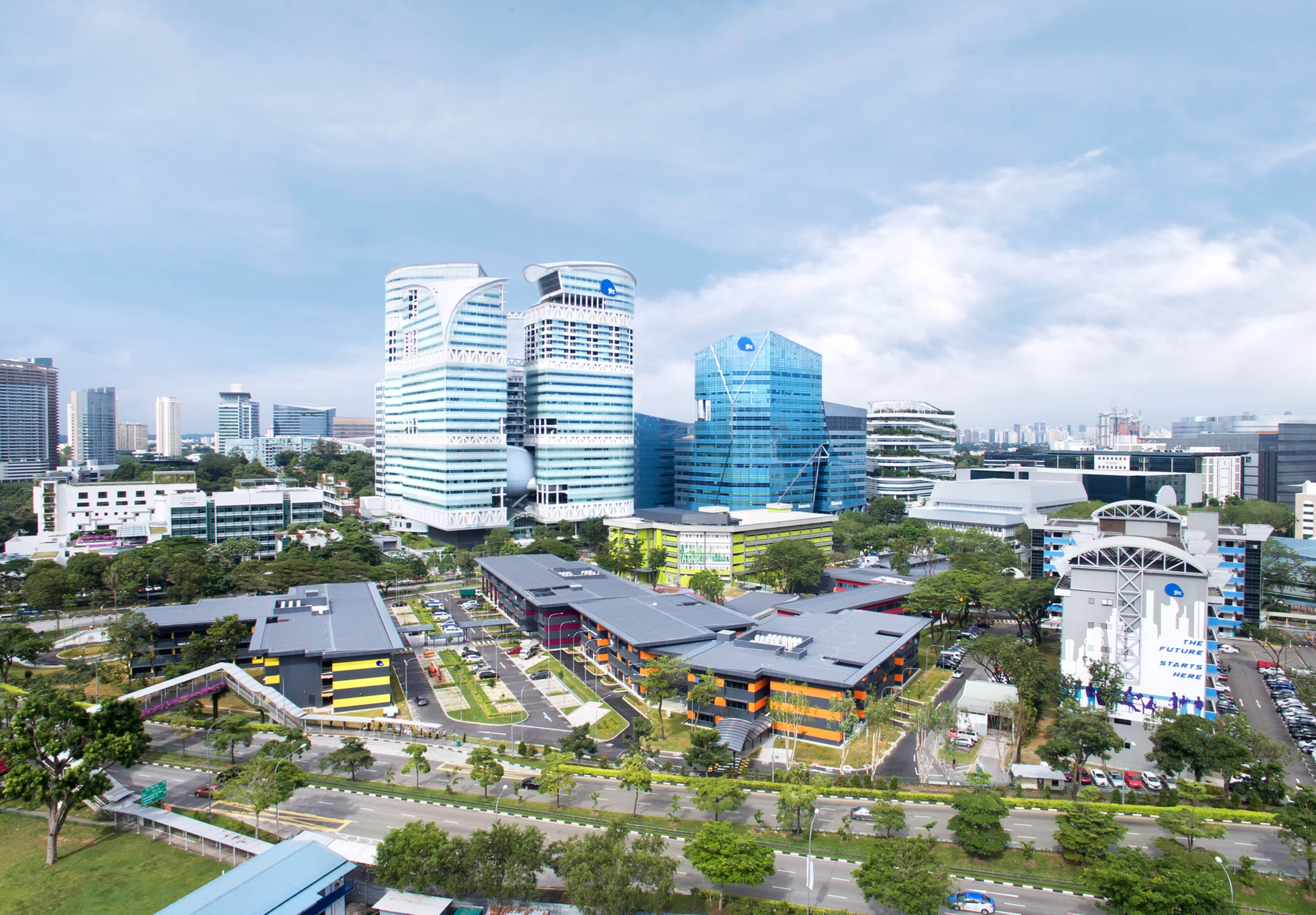 Aerial view of a modern urban area with tall buildings, a colorful complex, and surrounding greenery under a clear sky.