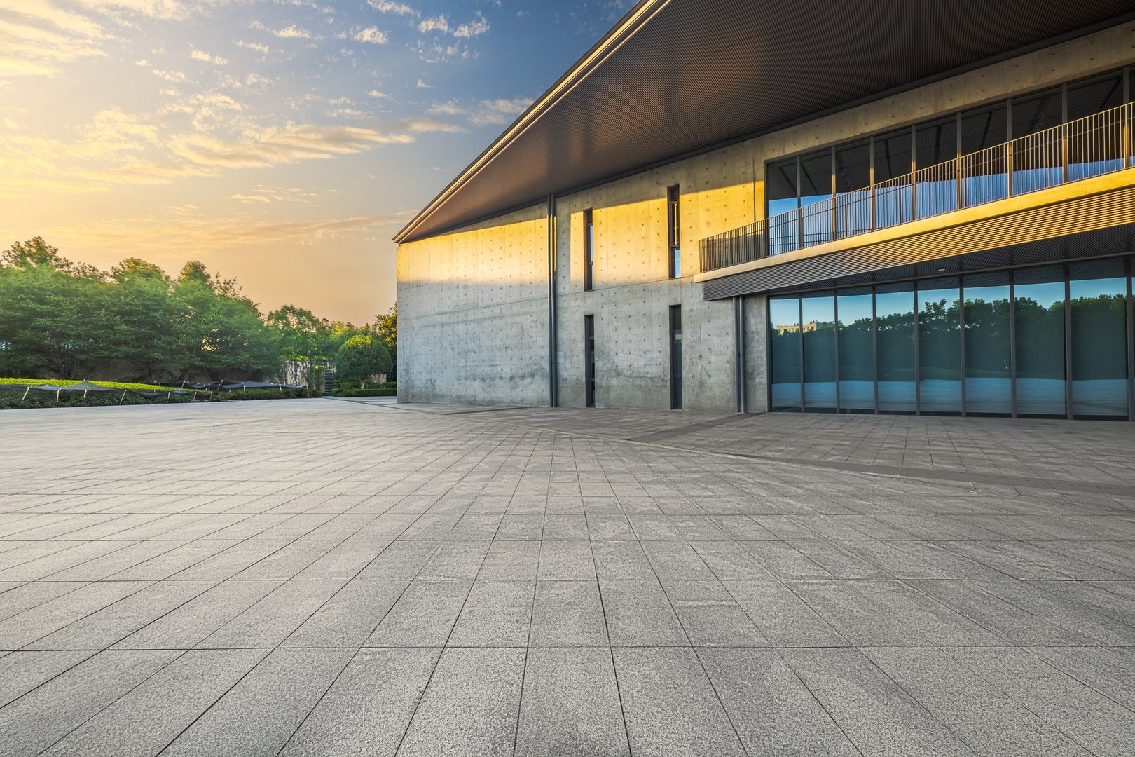 Empty stone paved square floor leads towards a modern concrete building exterior featuring large windows reflecting the early morning sunrise.