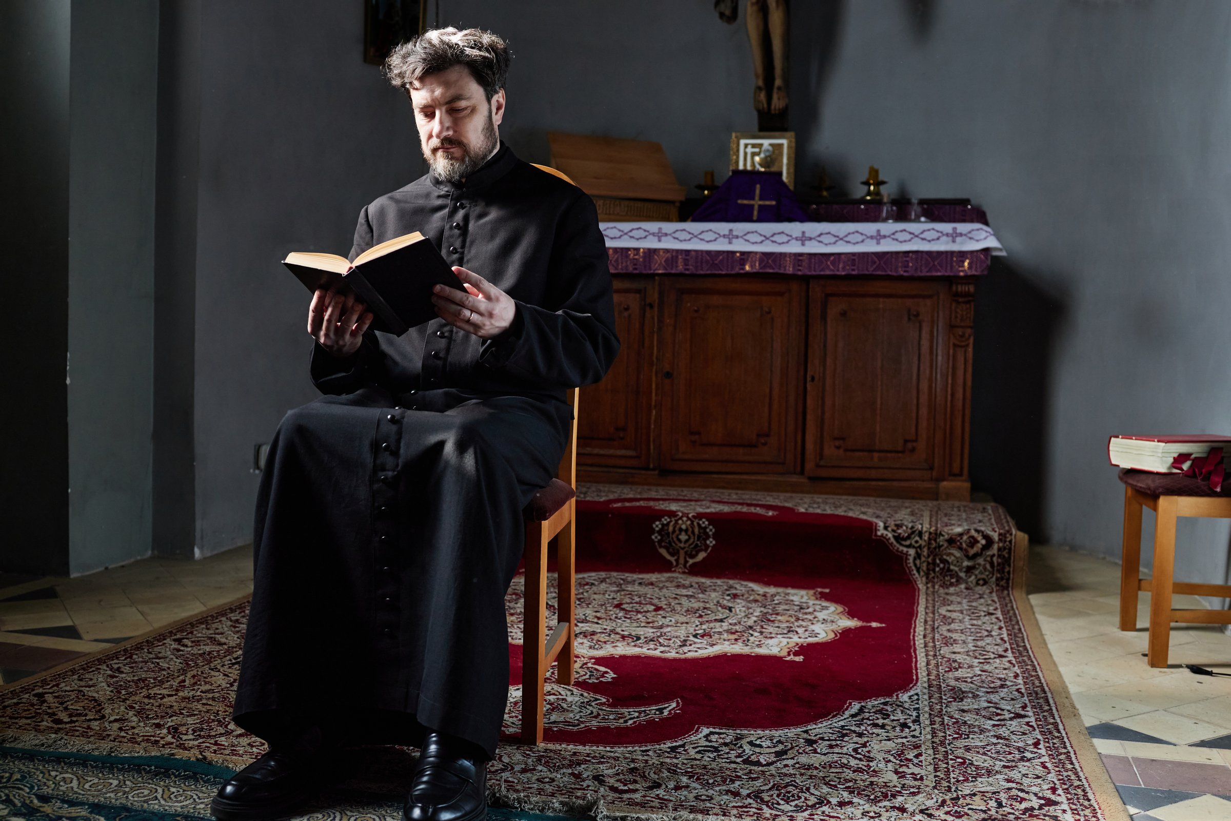 Middle aged Caucasian man wearing clerical robe sitting on chair in church reading book with serious expression, altar and religious objects visible in background