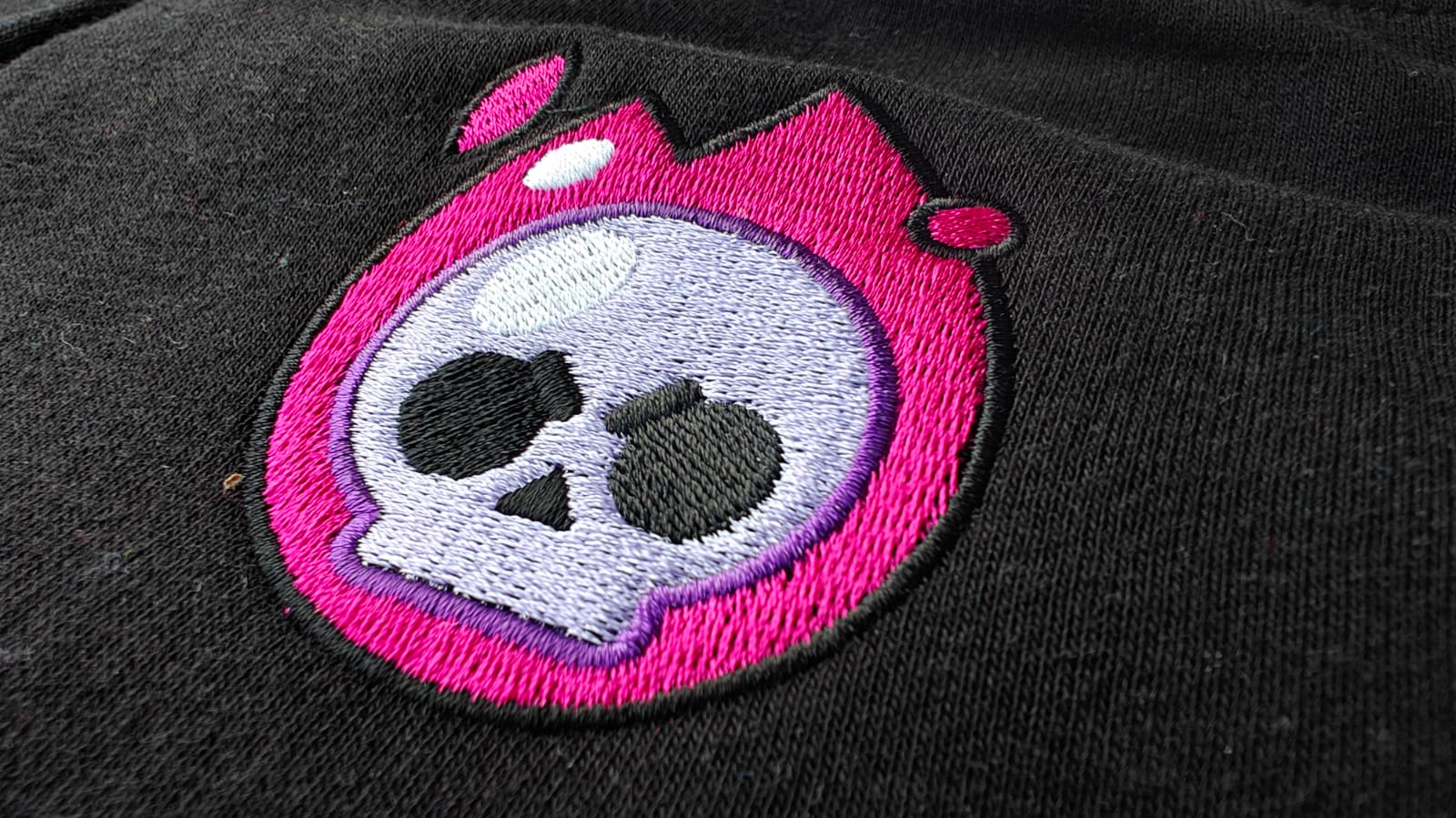 Close-up of a pink and purple embroidered skull logo on black fabric, featuring a flame-like design around the skull.