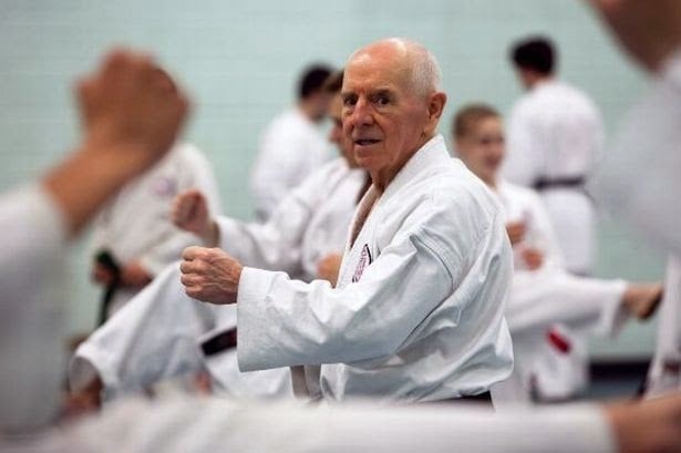 An older man practicing karate in a dojo with other participants visible in the background.
