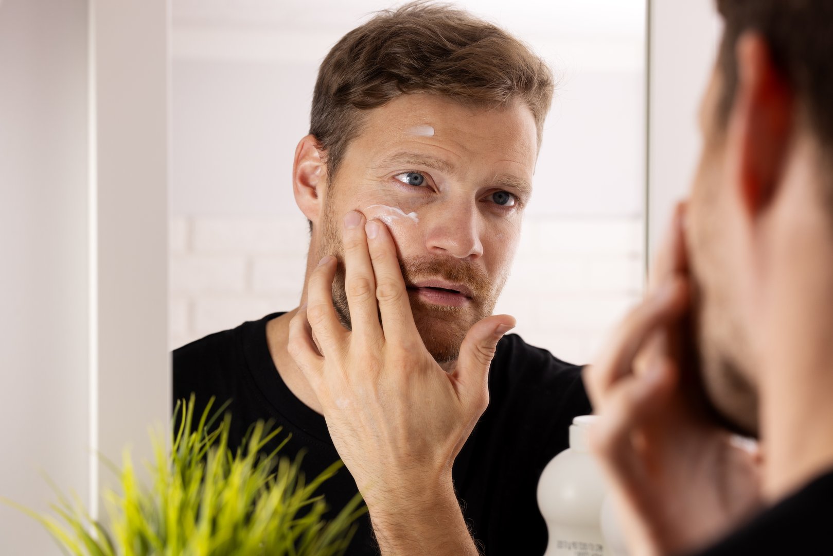 man applying facial cream in front of mirror at bathroom. male face skin care