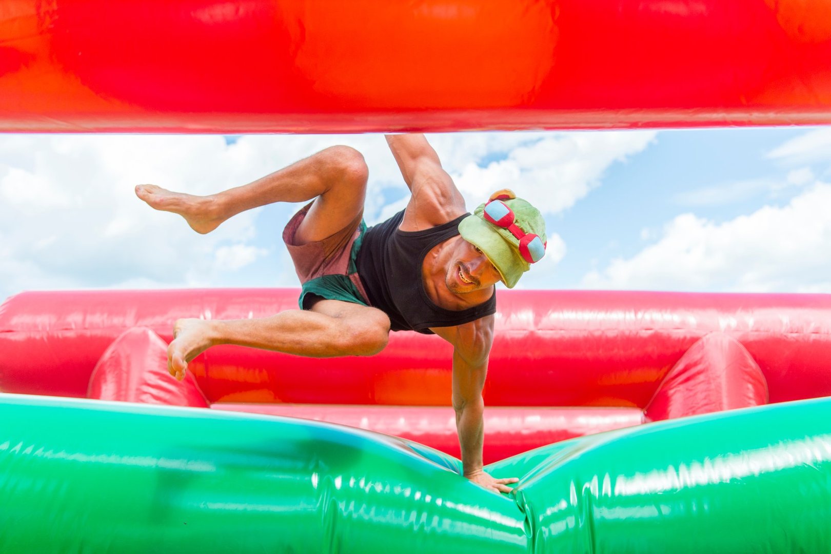 Man navigating an inflatable obstacle course outdoors, wearing a cap and sunglasses, with bright red and green sections.