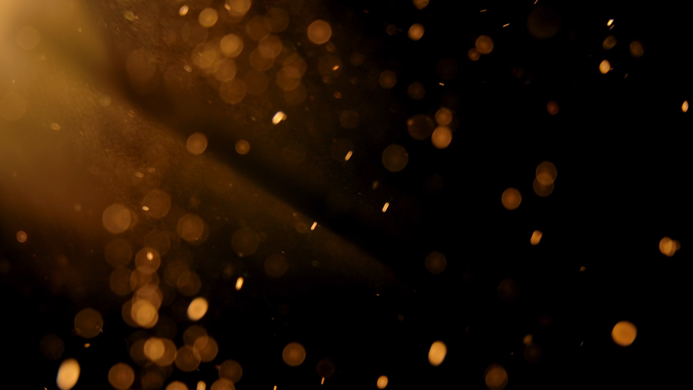Floating golden dust particles illuminated by a warm light beam in a dark space, creating a dreamy and atmospheric visual effect.
