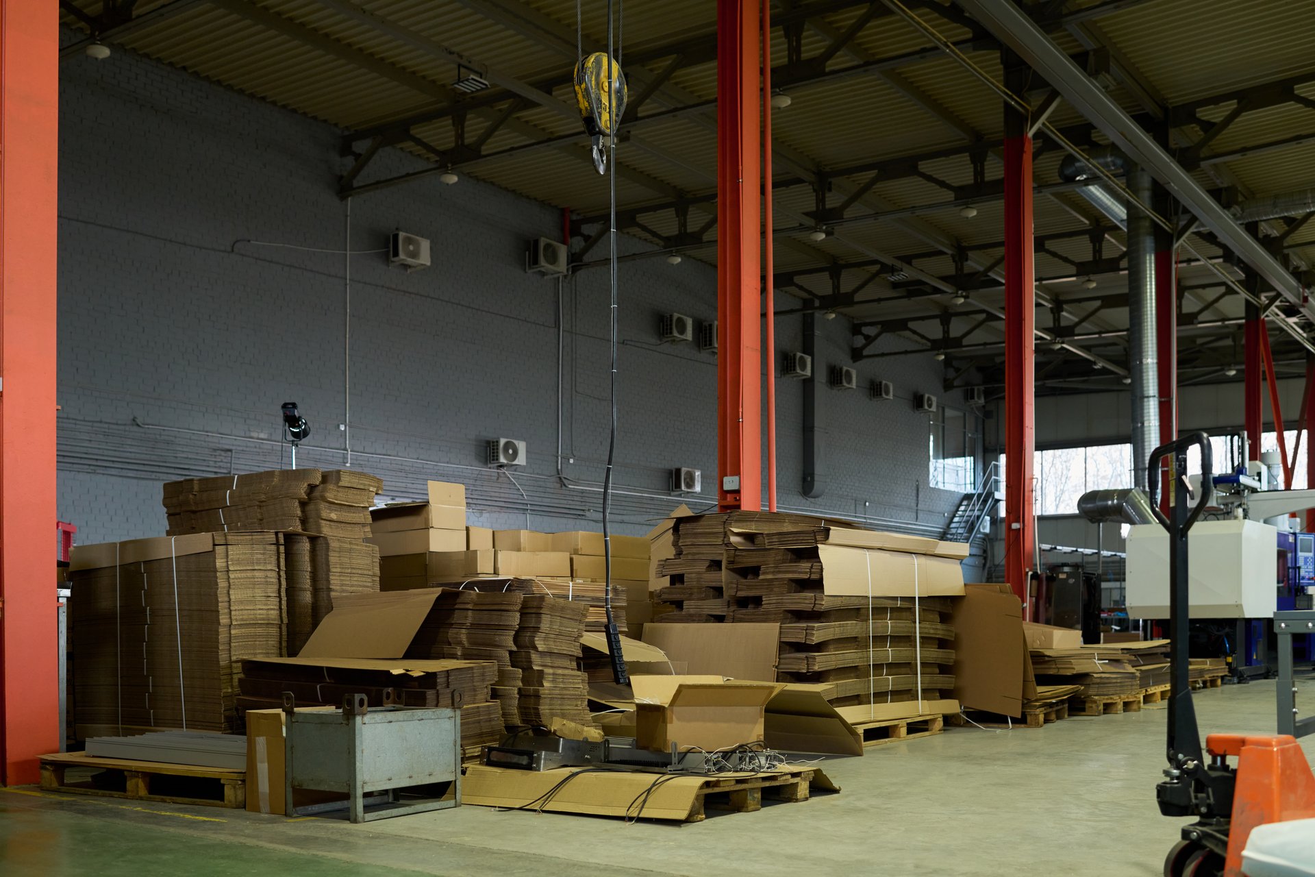 Row of stacked cardboard sheets on pallets and packed boxes standing on the floor of spacious warehouse with equipment