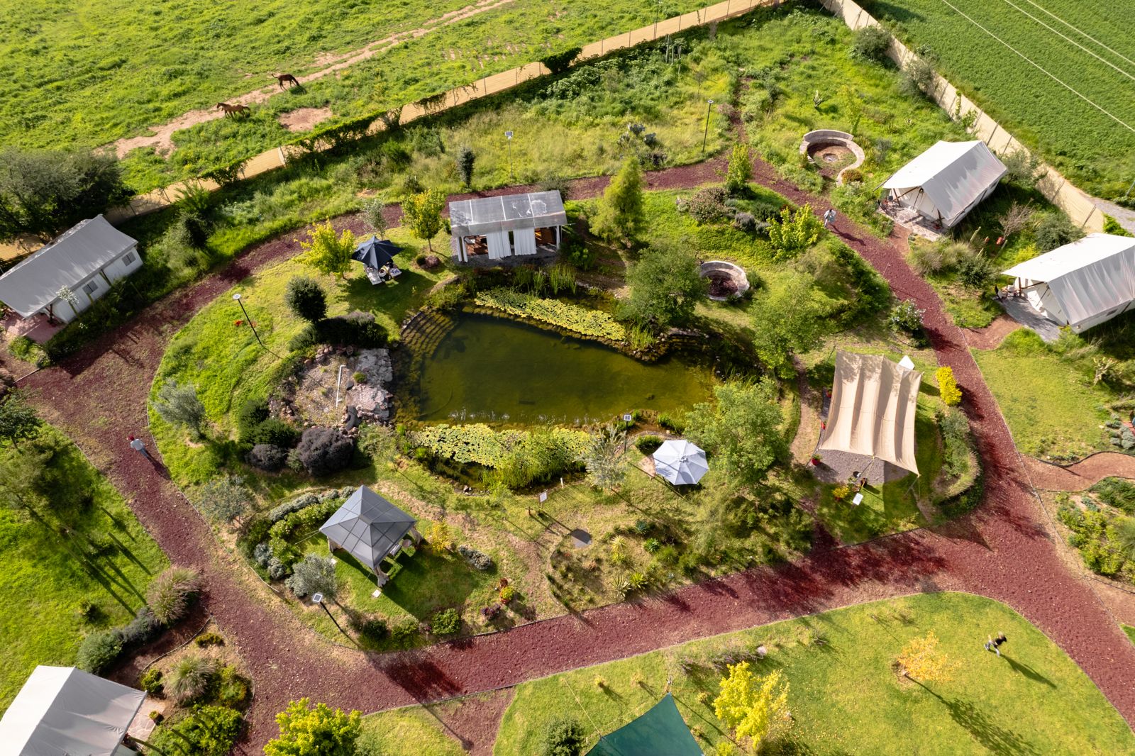 Aerial view of a landscaped garden with a pond, pathways, tents, and greenery in a rural setting.