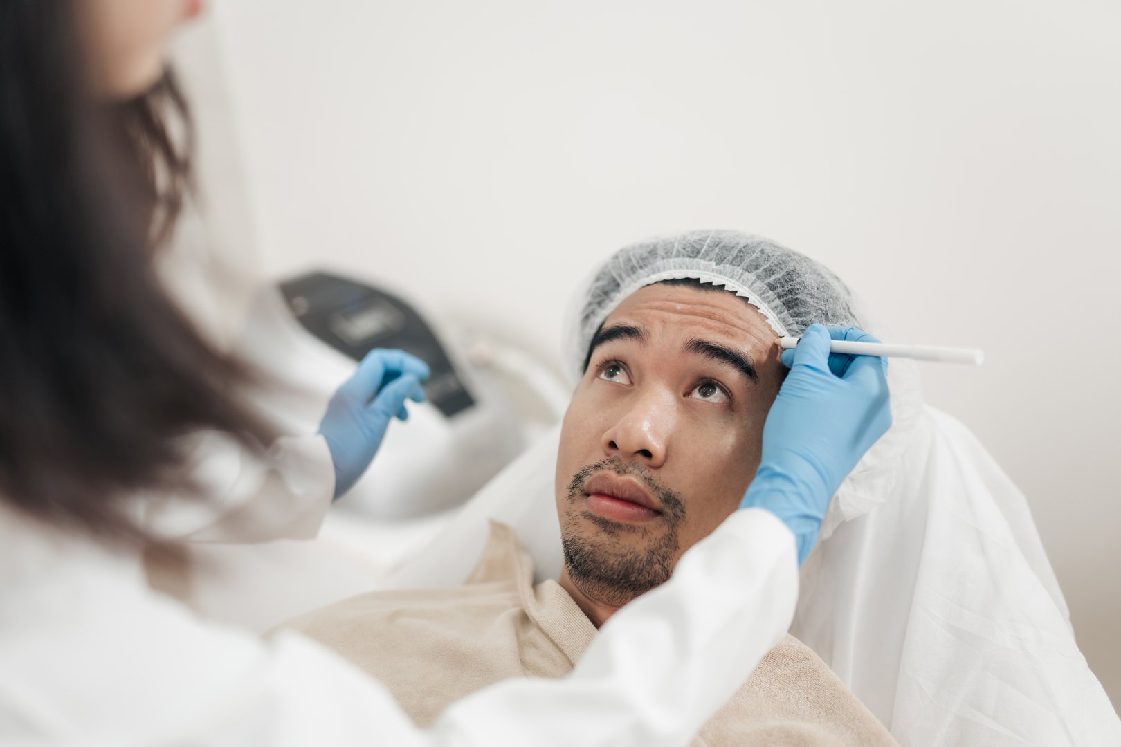 Anti aging injection on face. Asian man receiving pre-procedure markings on his forehead with a pen by face care specialist.