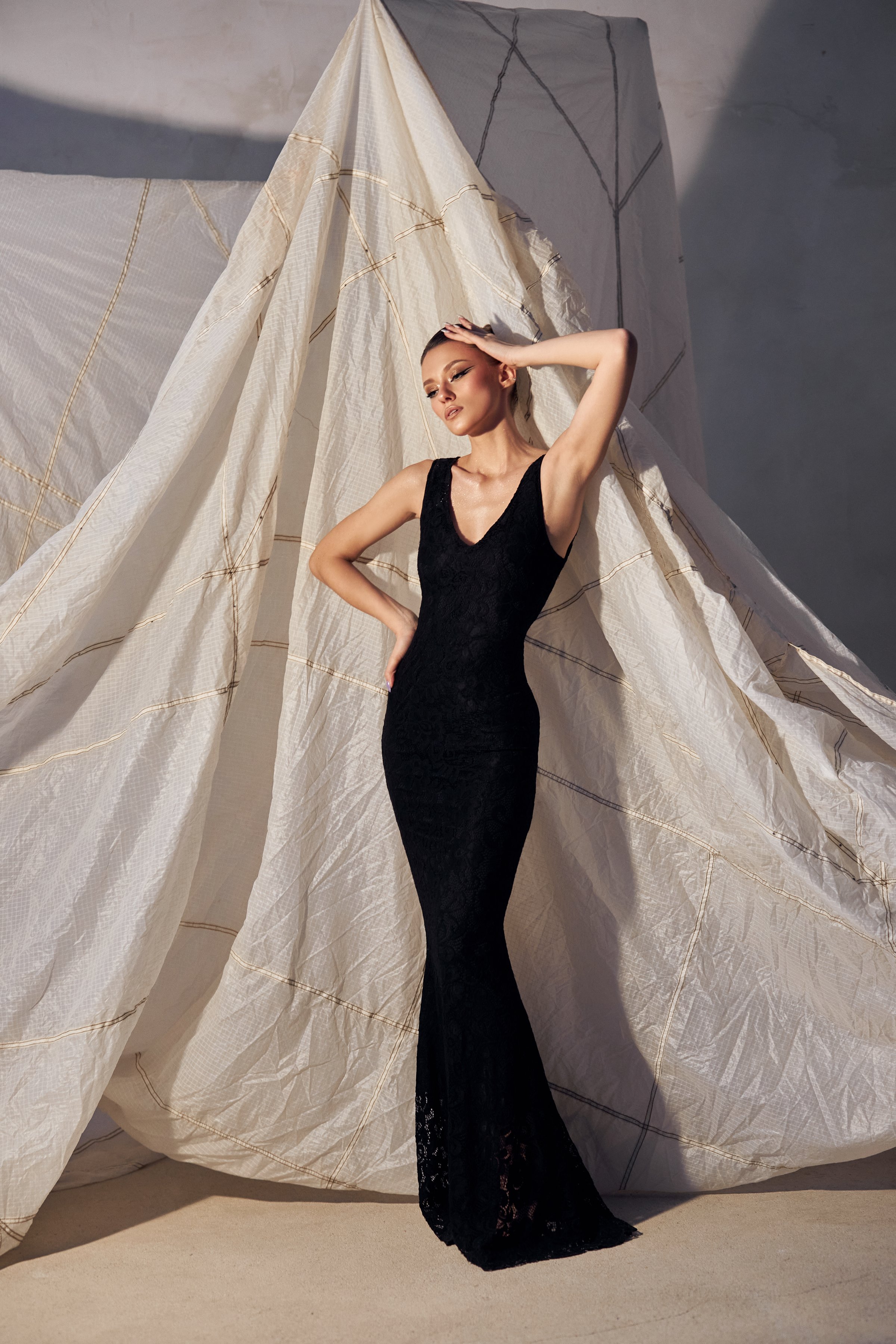 woman stands elegantly in a fitted black evening gown, showcasing her poise. backdrop features draped white fabric, creating a stylish and minimalist atmosphere in the studio.