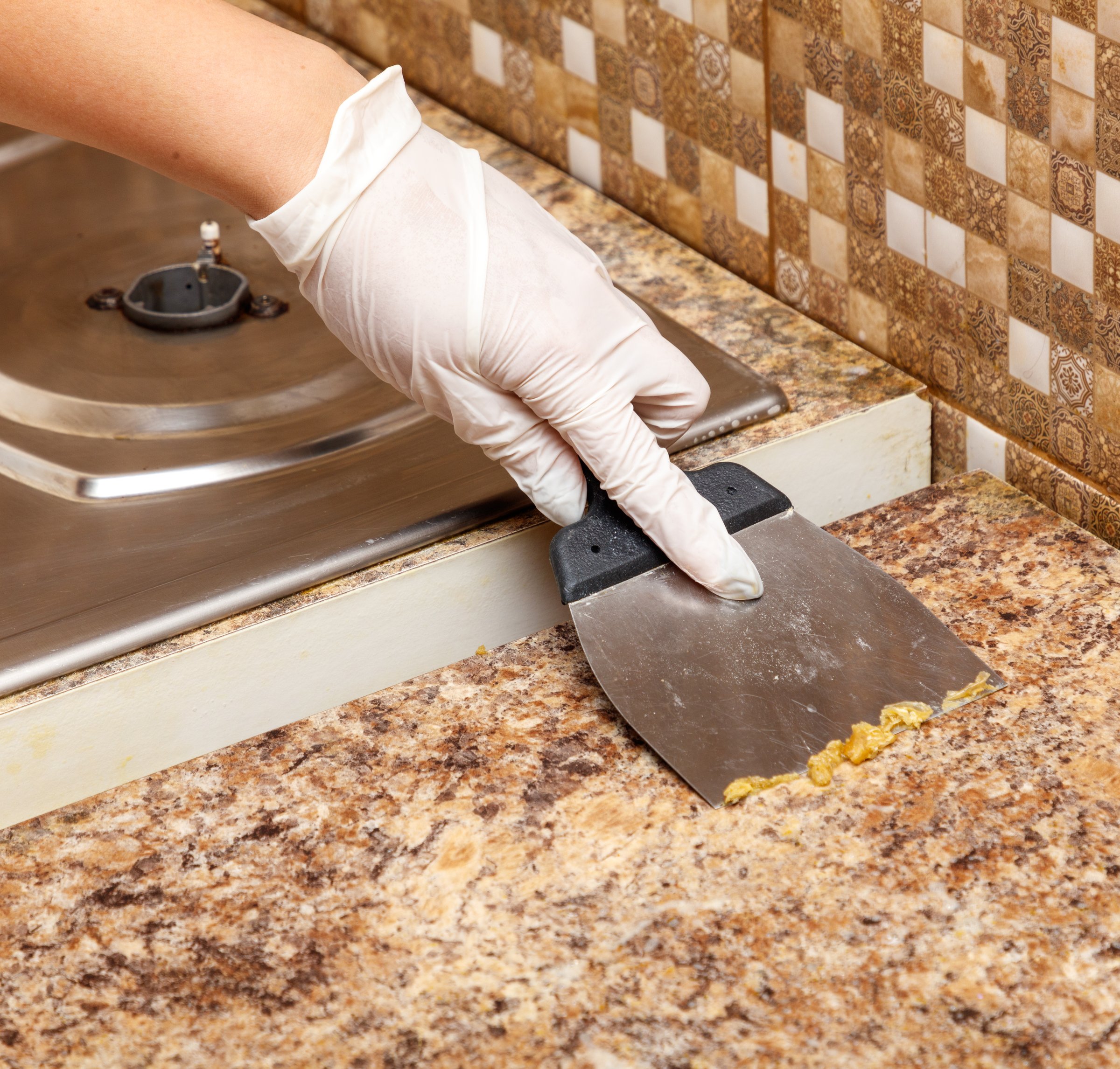 A person is using a spatula to spread something on a countertop. The countertop is made of granite and has a checkered pattern. The person is wearing gloves while using the spatula