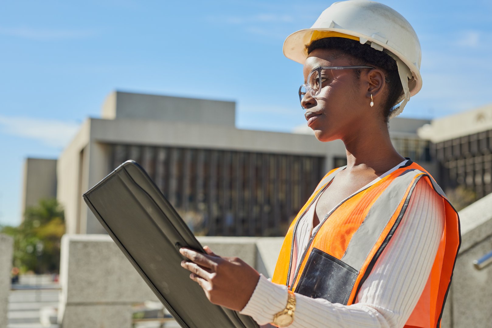 A professional female engineer wearing a hard hat and safety vest stands at a construction site, holding a portfolio. She appears focused and confident as she evaluates the urban project around her. The image captures the essence of dedication and expertise in modern construction and engineering fields.