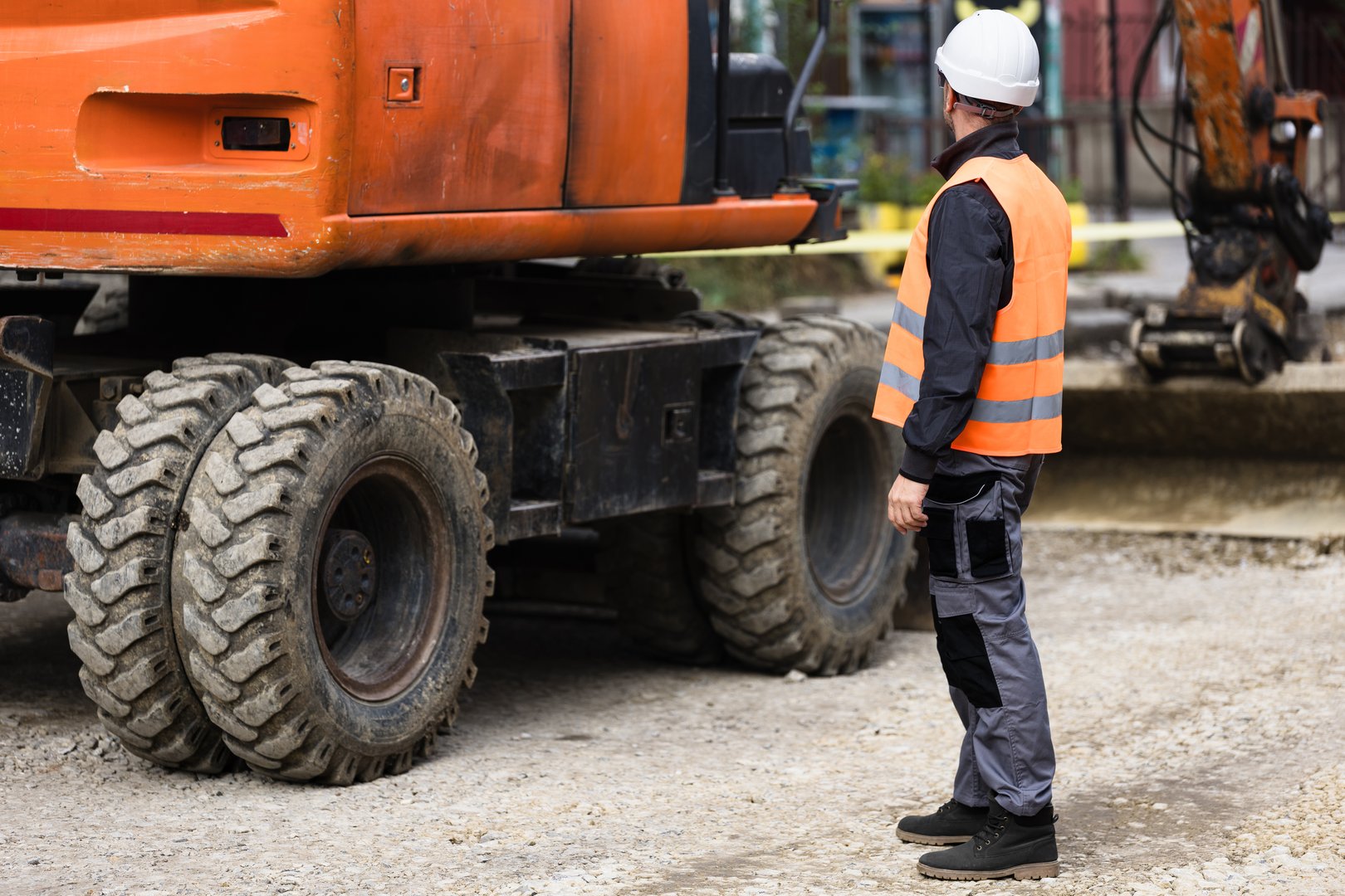 A construction worker wearing a safety vest and helmet closely examines a large piece of machinery at a busy construction site in daylight.