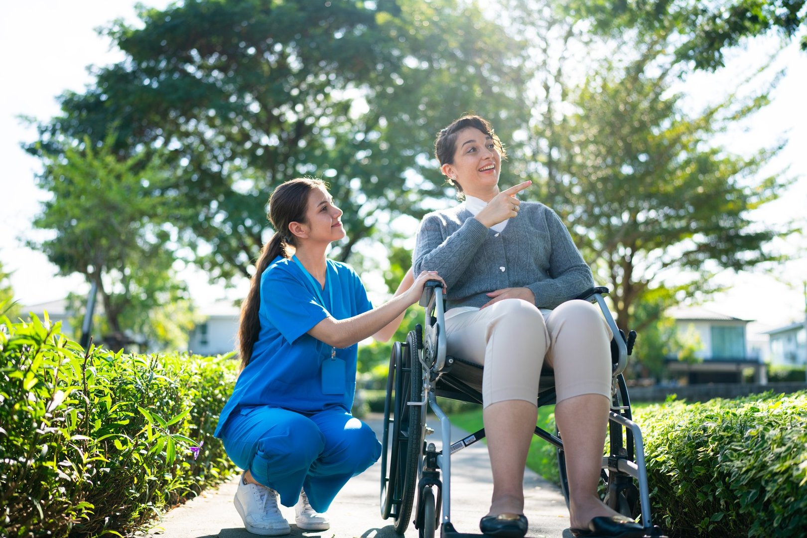 A nurse in blue scrubs kneeling beside a woman in a wheelchair, engaging in conversation while enjoying a bright, sunny park setting.
