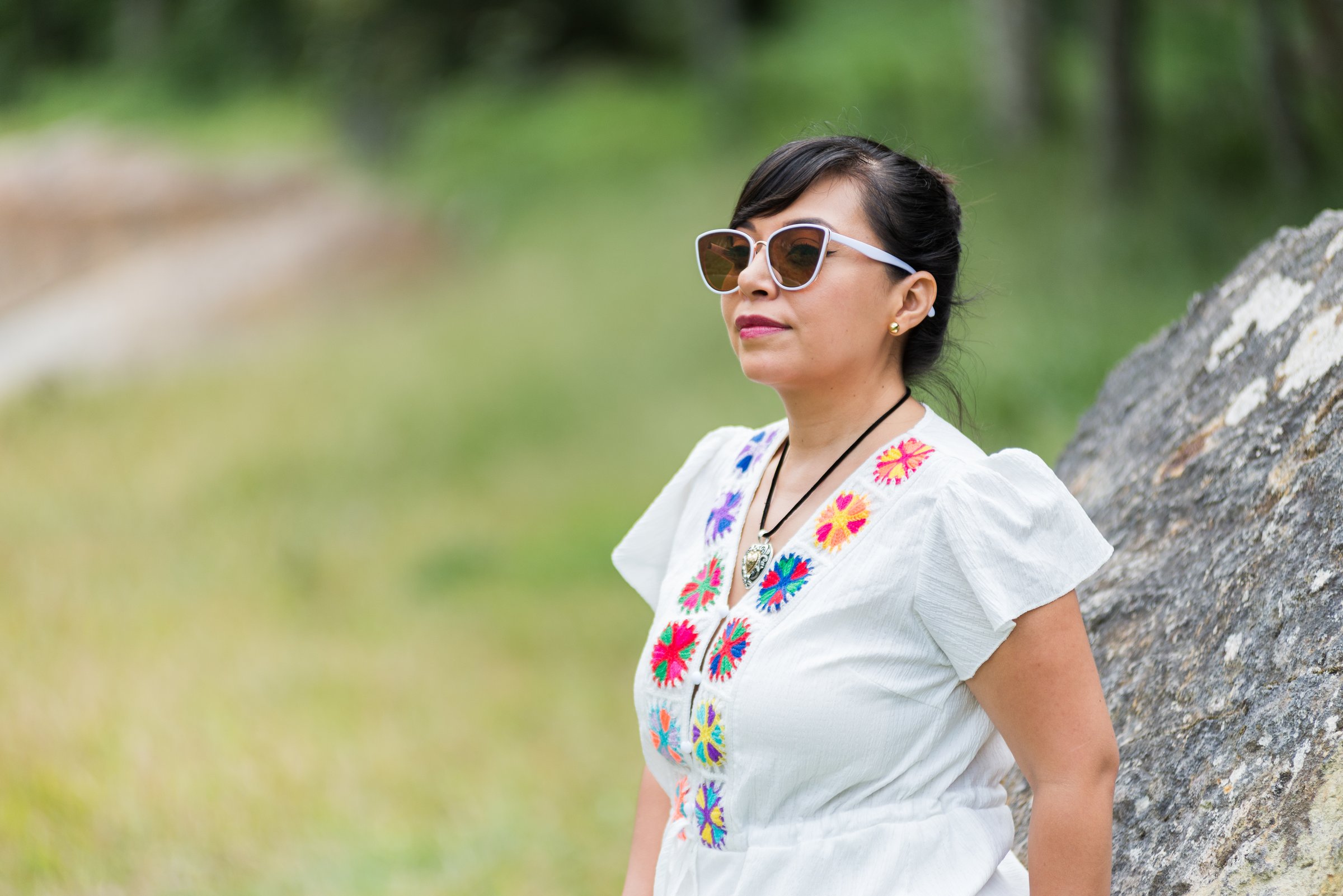 A woman with dark hair, wearing sunglasses and a white blouse adorned with colorful embroidery, stands outdoors near a rock.