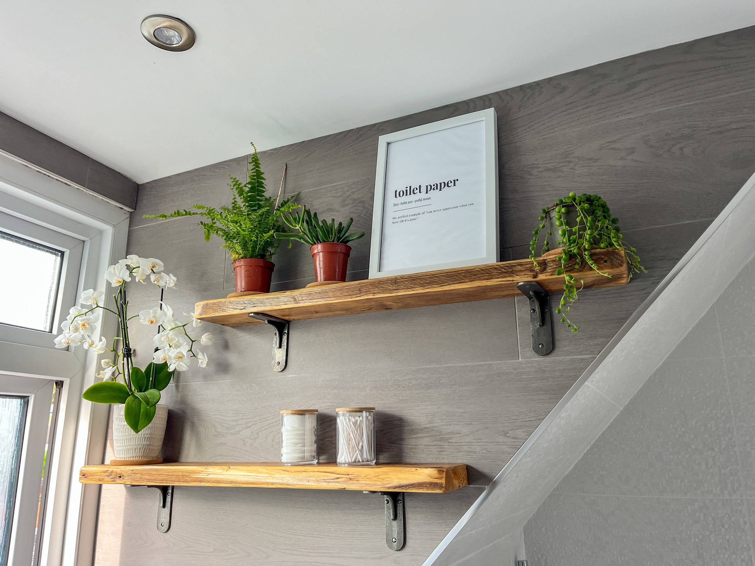 Natural wooden shelves against a grey, tiled wall. There is a fern and succulents on the shelves