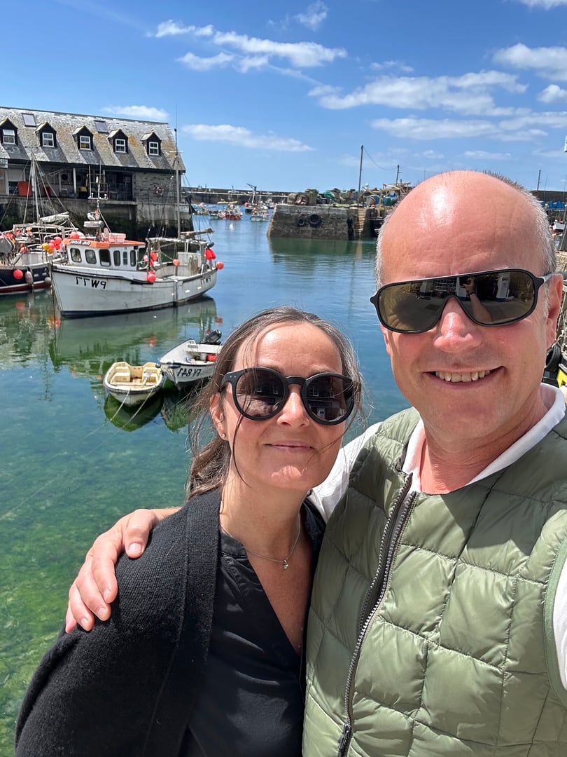 Smiling couple wearing sunglasses posing by a scenic harbor with fishing boats and a sunny sky in the background.
