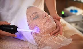 Person receiving a facial treatment with a high-frequency wand glowing purple under a gauze mask.