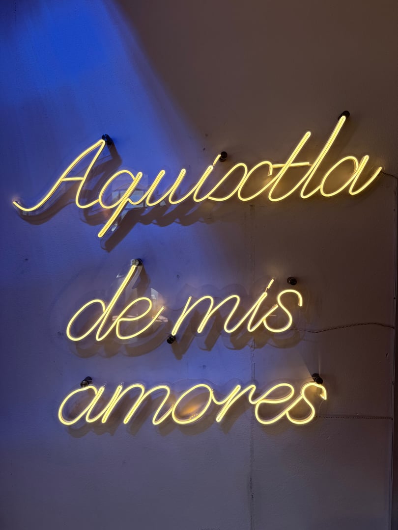 Yellow neon sign reading "Aquixtla de mis amores" on a textured wall with a blue light casting a glow on one side.