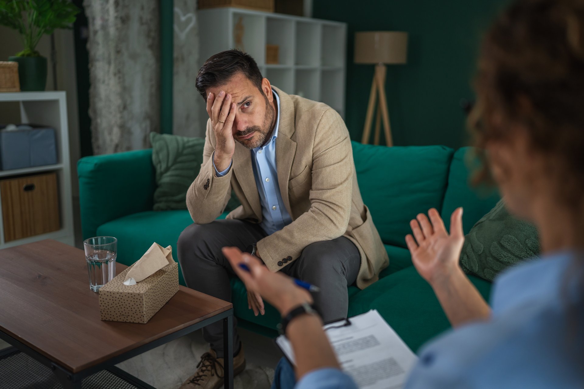 Man covering his face with hand, struggling with emotional pain and mental health issues while receiving support from a therapist in a counseling session