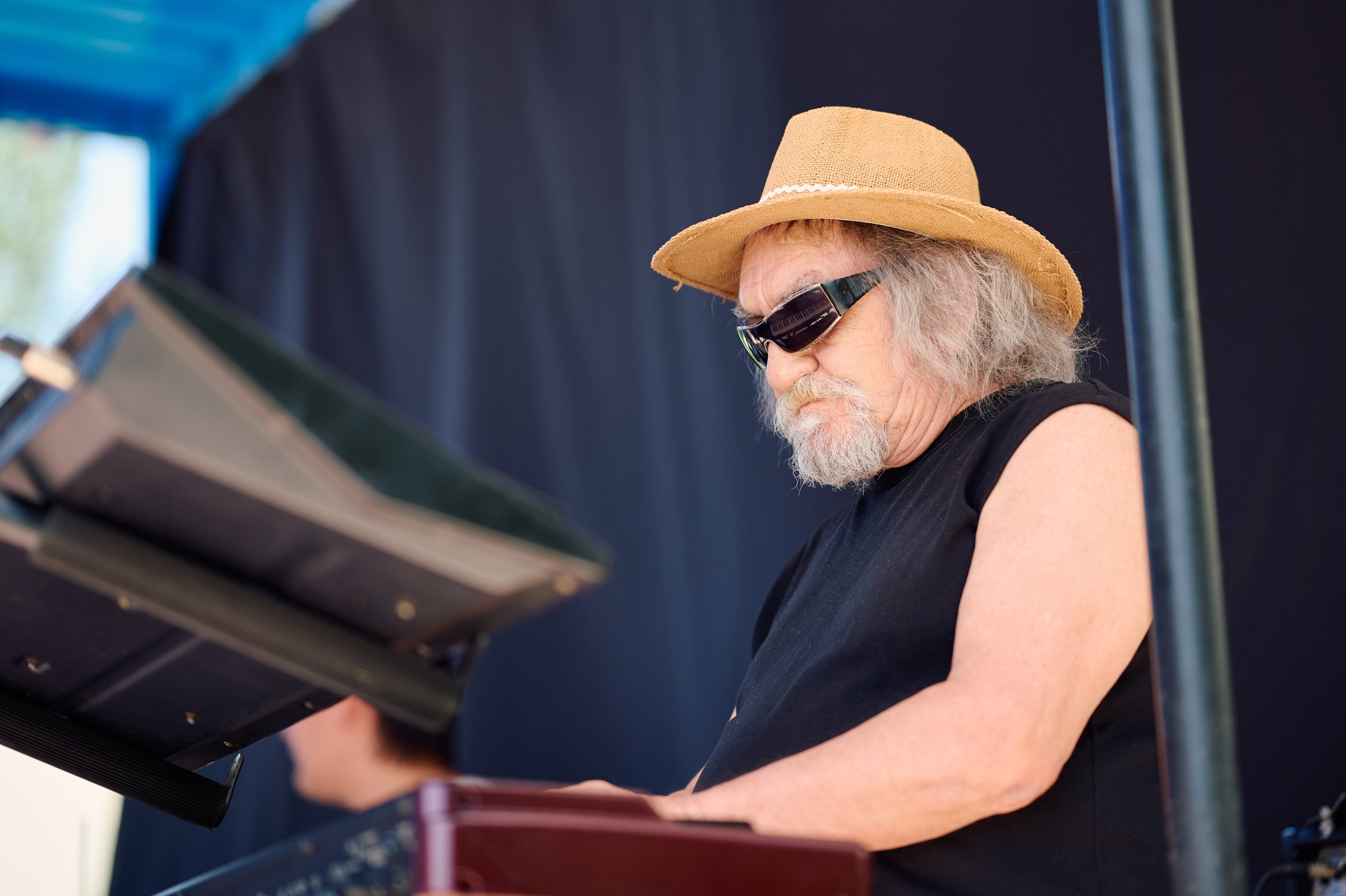 Senior musician playing keyboard at an outdoor concert, sporting sunglasses and a hat while captivating the audience with vibrant melodies