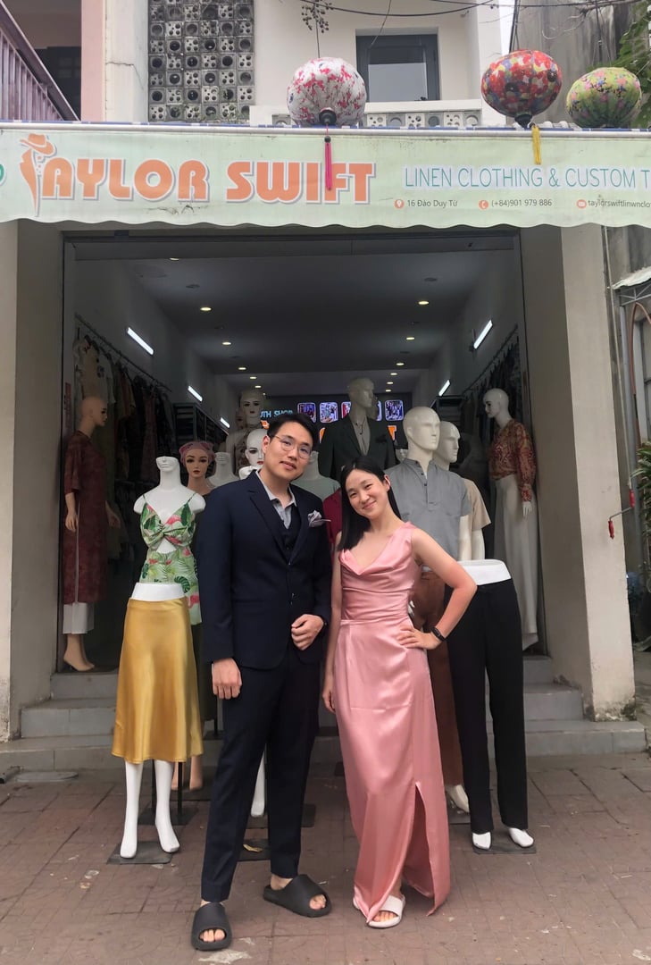 Two people pose in front of a clothing shop named "Taylor Swift" with mannequins displaying various outfits.