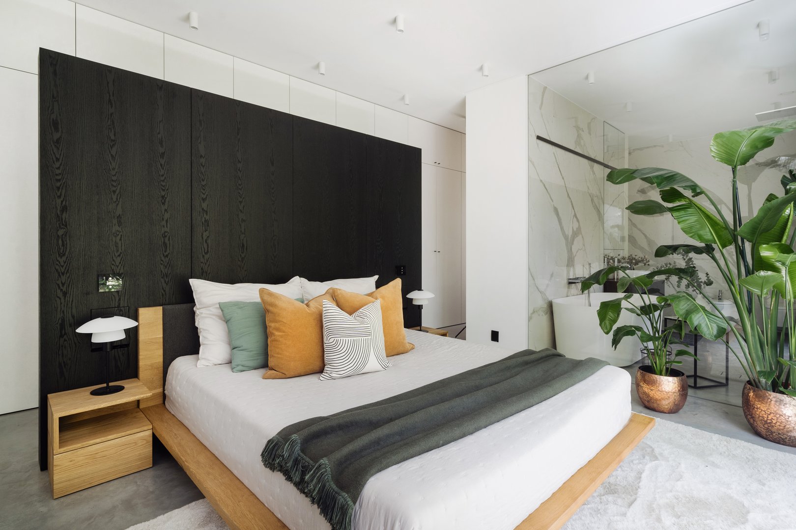 A contemporary bedroom showcases a double or king size bed with decorative pillows and a cozy throw, complemented by large indoor plants and minimalist design elements. Room with bathroom