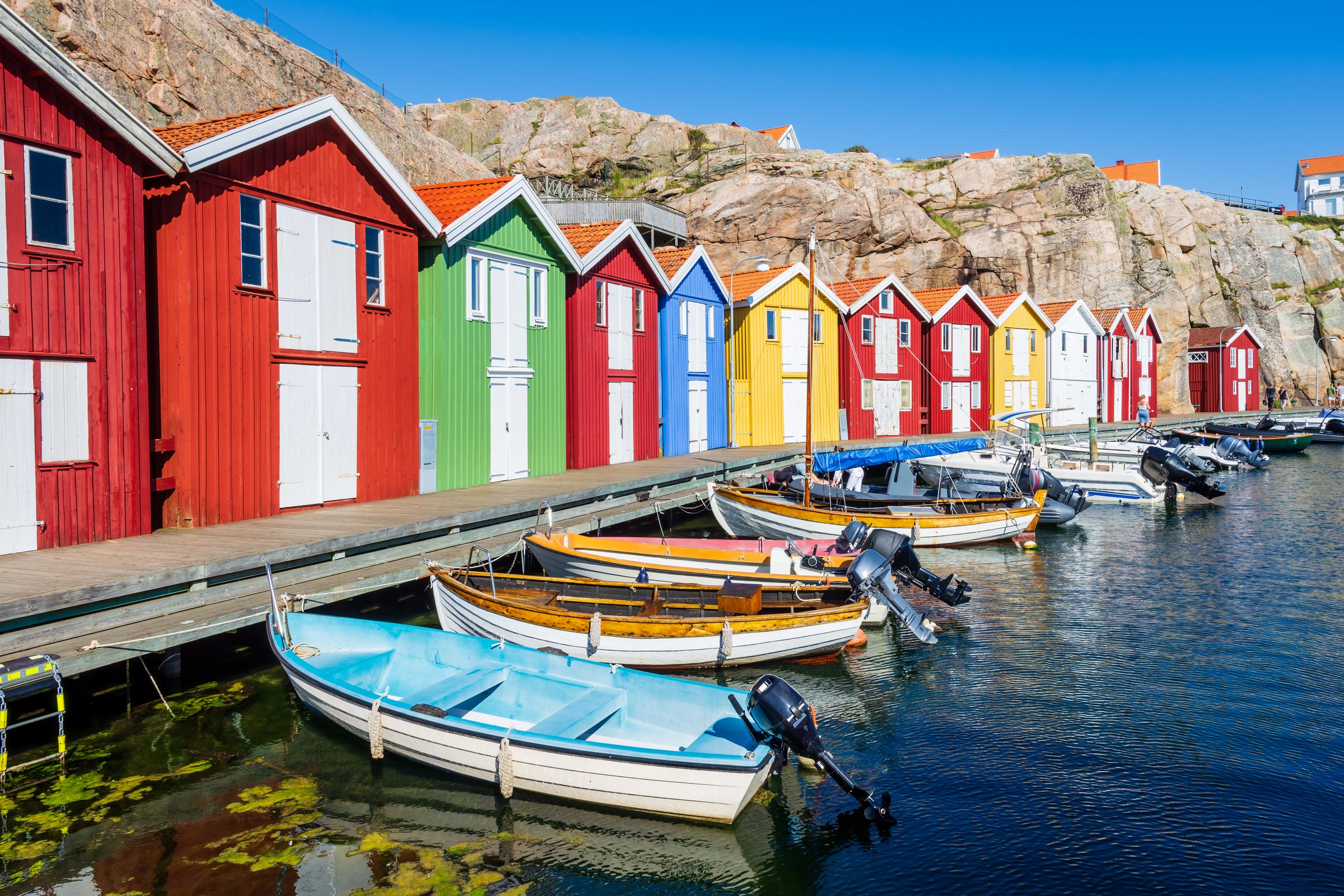 Colorful wooden boathouses lined up against the rocky cliff in the old fishing harbor of Smögen, Sweden, with boats moored to the boardwalk on a sunny summer day.