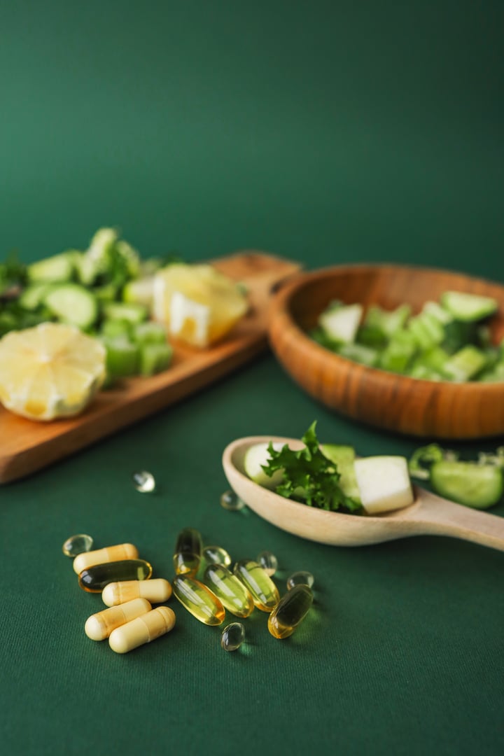 Overhead view of chopped celery, cucumber, and lemon alongside vitamin capsules on a wooden board against a green background. Perfect for healthy lifestyle or detox meal prep.