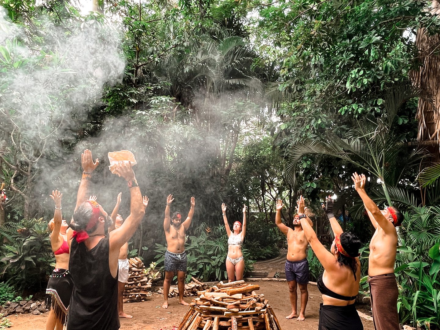 Group performing a spiritual ritual with hands raised around a fire pit in a tropical forest setting.
