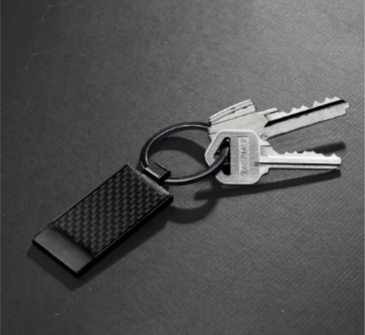 A set of keys on a ring with a carbon fiber texturized fob on a dark surface.