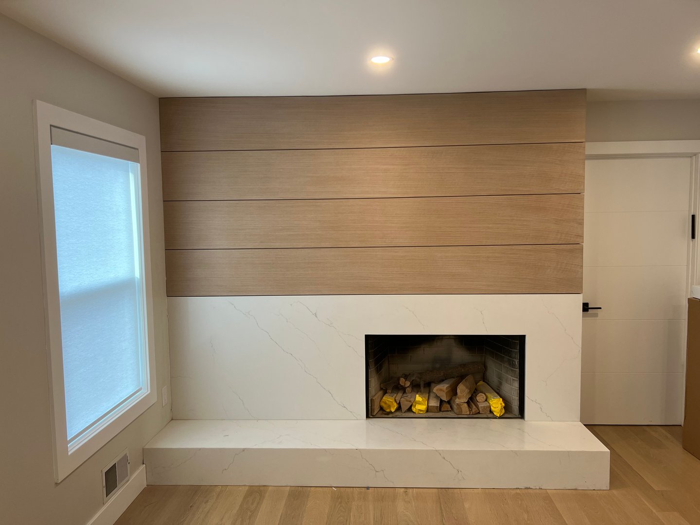 Modern living room with a sleek fireplace, marble surround, wood paneling, and a large window with a white shade.