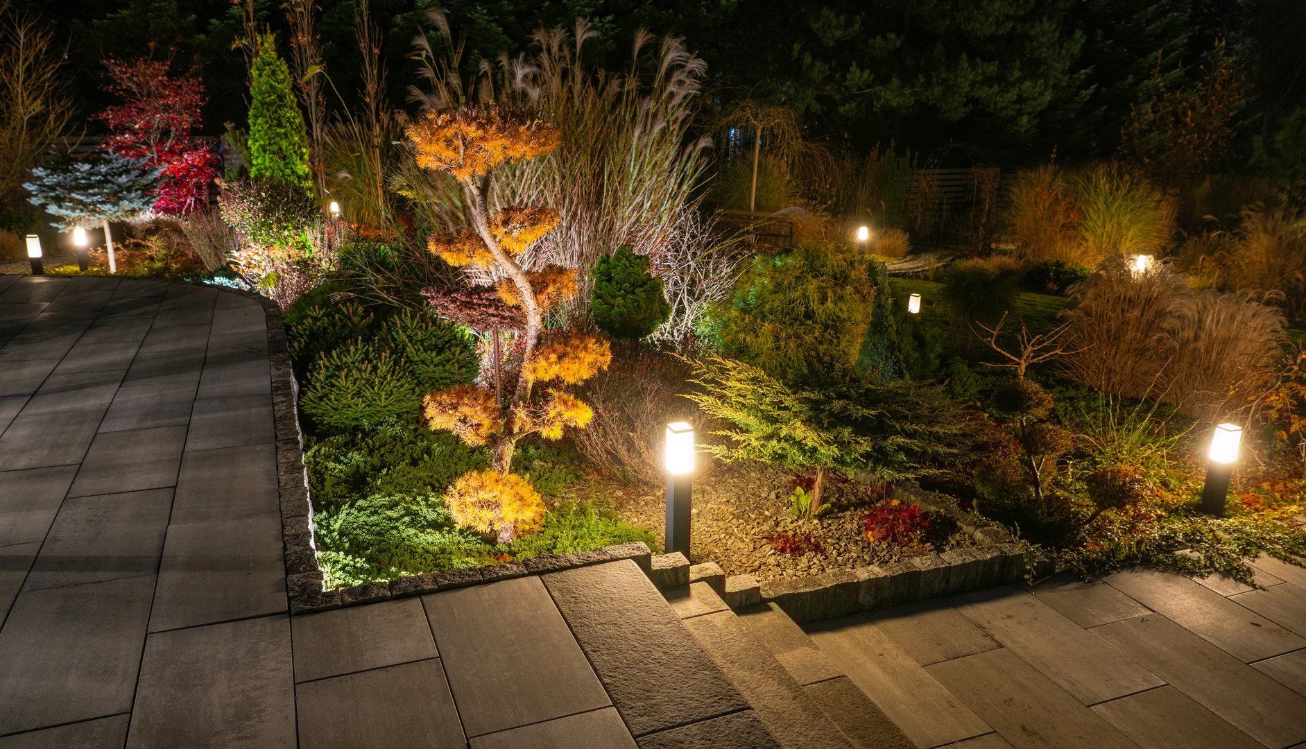 Warm lights illuminate the garden pathway, highlighting vibrant trees and manicured plants, creating a serene atmosphere for evening strolls.
