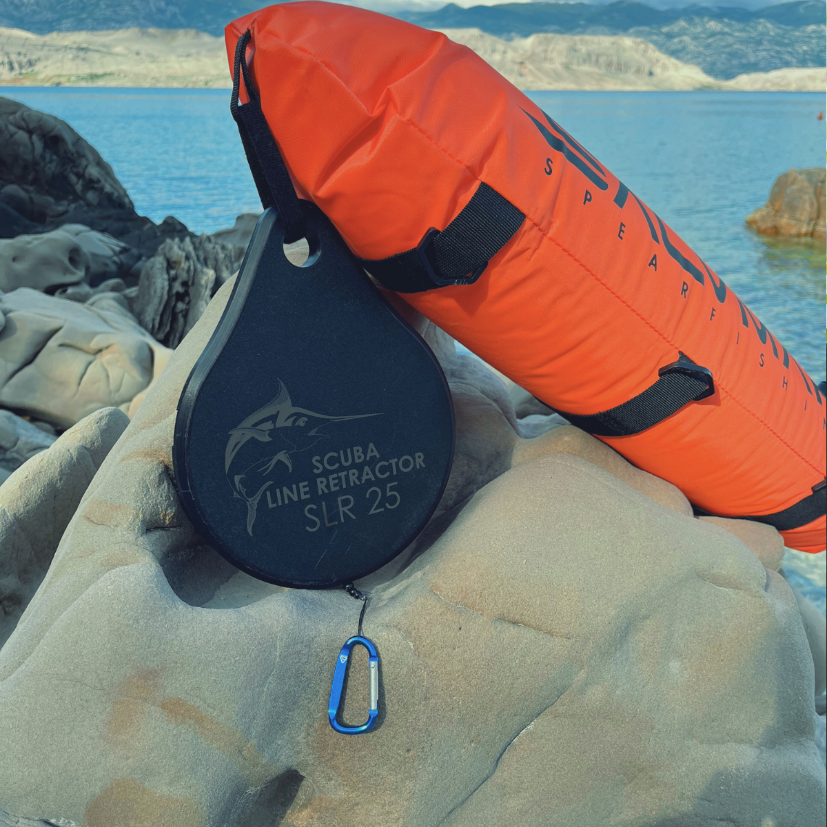 Scuba line retractor and orange float on rocky shore, with a view of the sea and distant mountains.