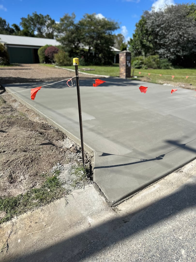 Freshly laid concrete driveway with safety tape, bordered by grass and trees, leads to a residential garage.