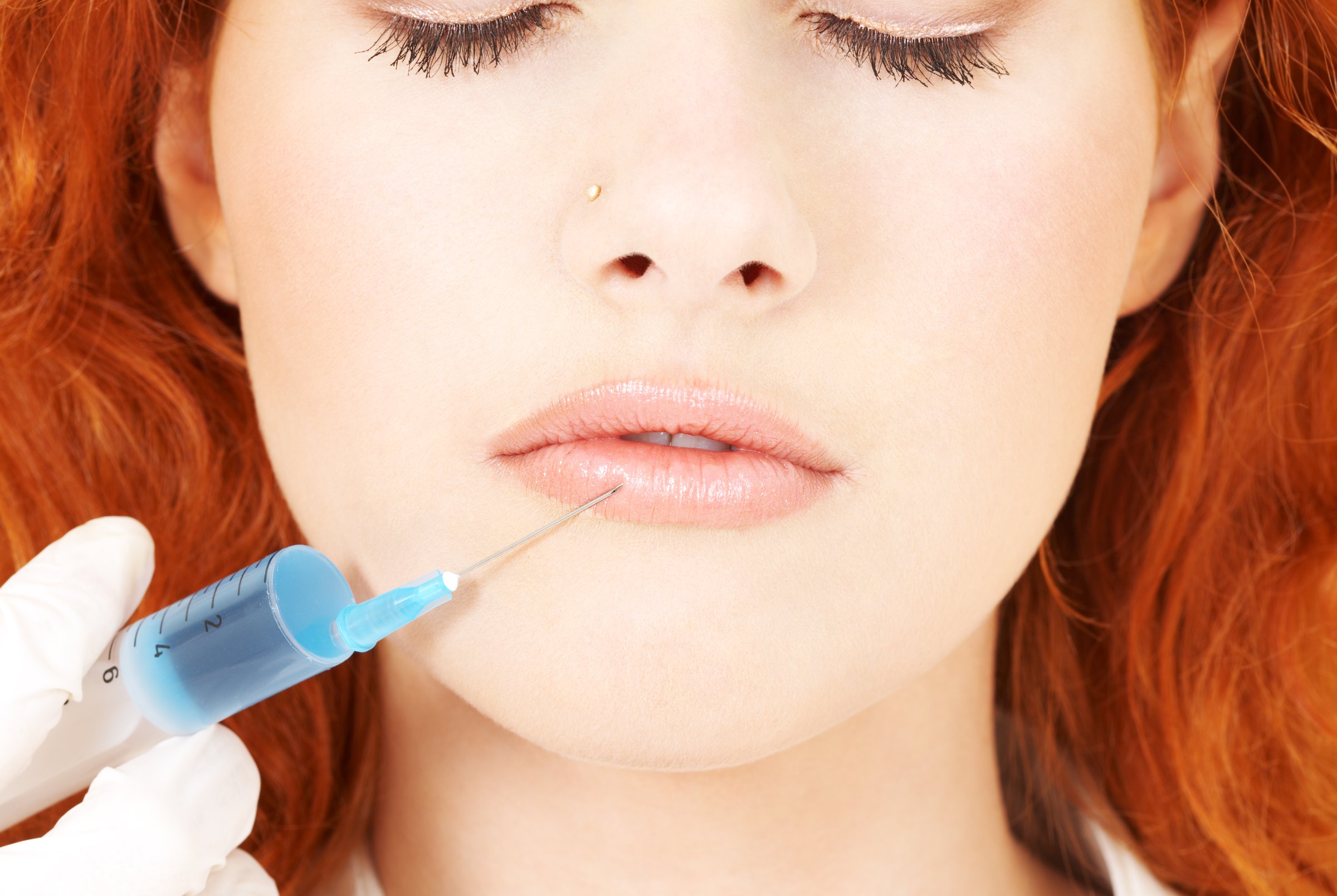 Close-up of a syringe near a womans lips, suggesting cosmetic injection. The woman has red hair and is wearing makeup.