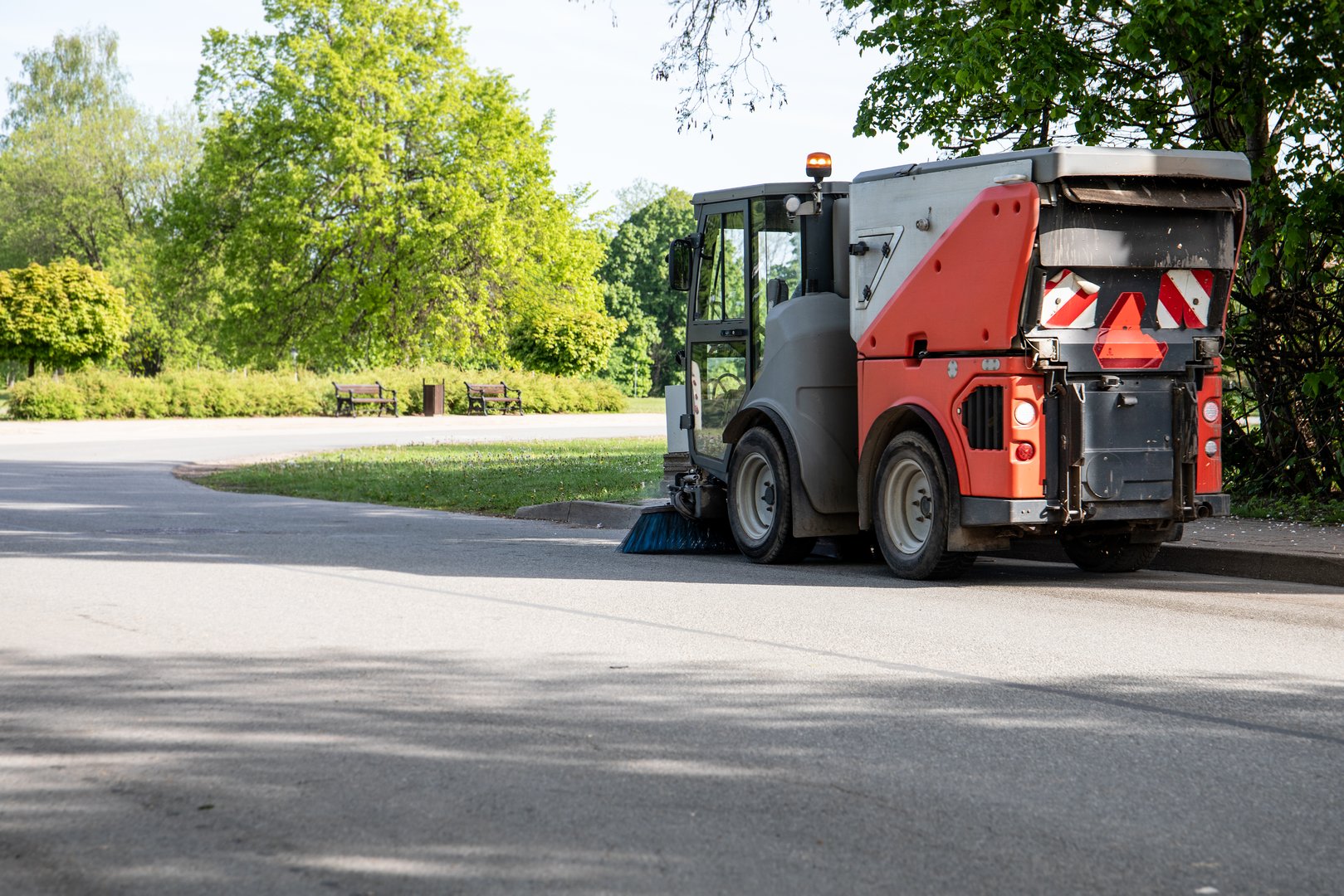 Street Sweeper Vehicle Cleaning Urban Roadside Near Park with Trees and Benches.