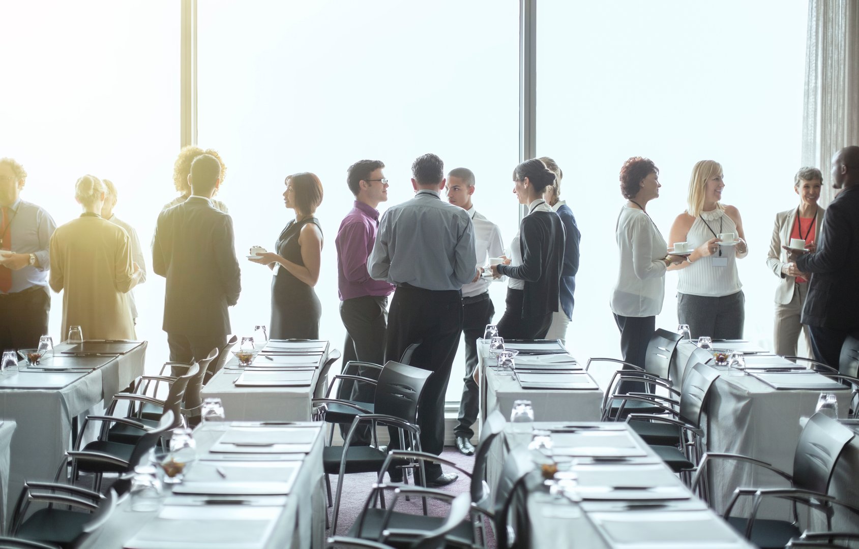 People networking in a bright conference room near large windows, with conference tables and chairs in the foreground.