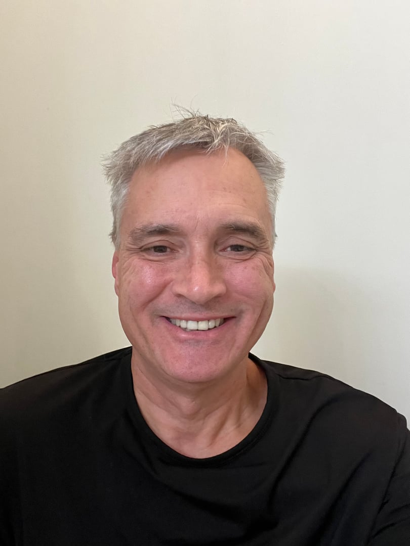 Man in a white shirt and tie smiles in front of a painting of sailboats on a sunny day.