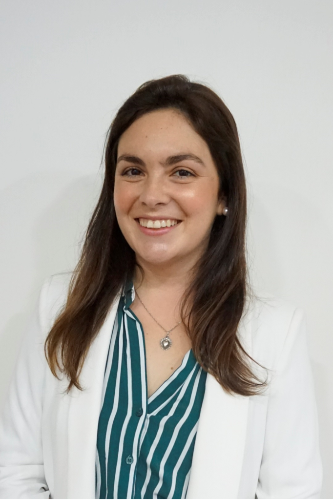 Woman smiling, wearing a white blazer over a striped shirt, with long brown hair against a plain white background.