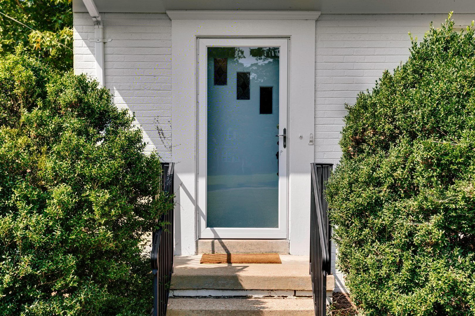 The entrance features a modern glass front door surrounded by green hedges, concrete steps, and metal railings, creating an inviting and tidy first impression.
