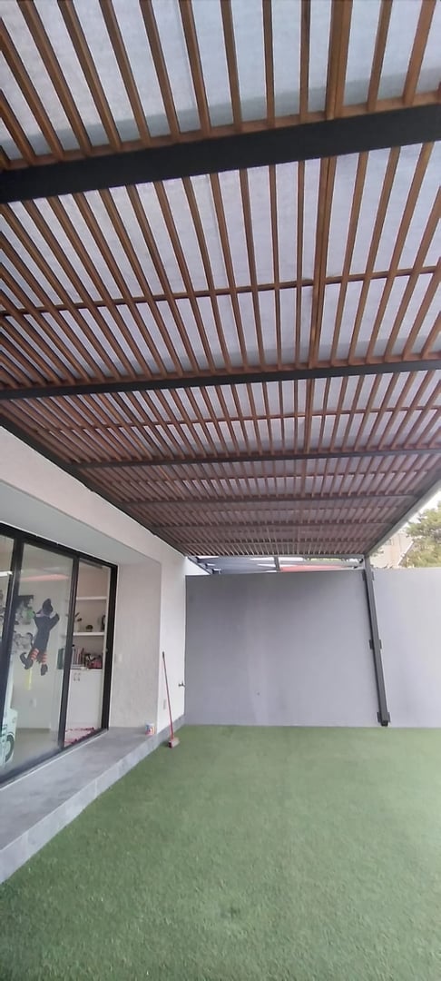 Modern pergola with wooden slats and a translucent roof over artificial turf in a backyard setting.