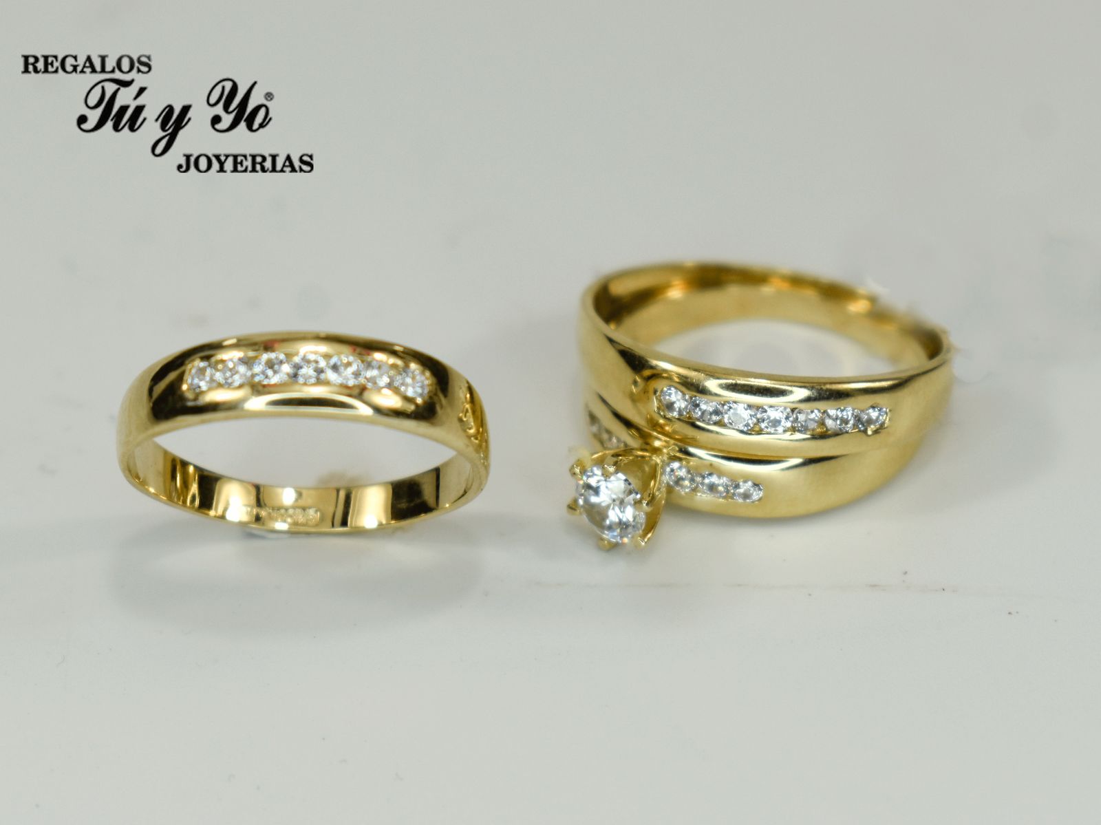 Two gold rings with embedded diamonds on a white surface, one with a prominent central diamond. Brand name "Tú y Yo Joyerías" visible.