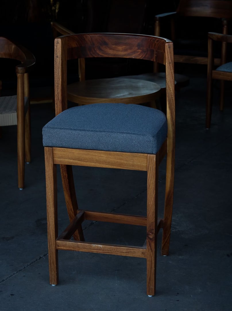 Banco alto de parota con respaldo bajo curvo envolvente y asiento tapizado en tela gris-azulada