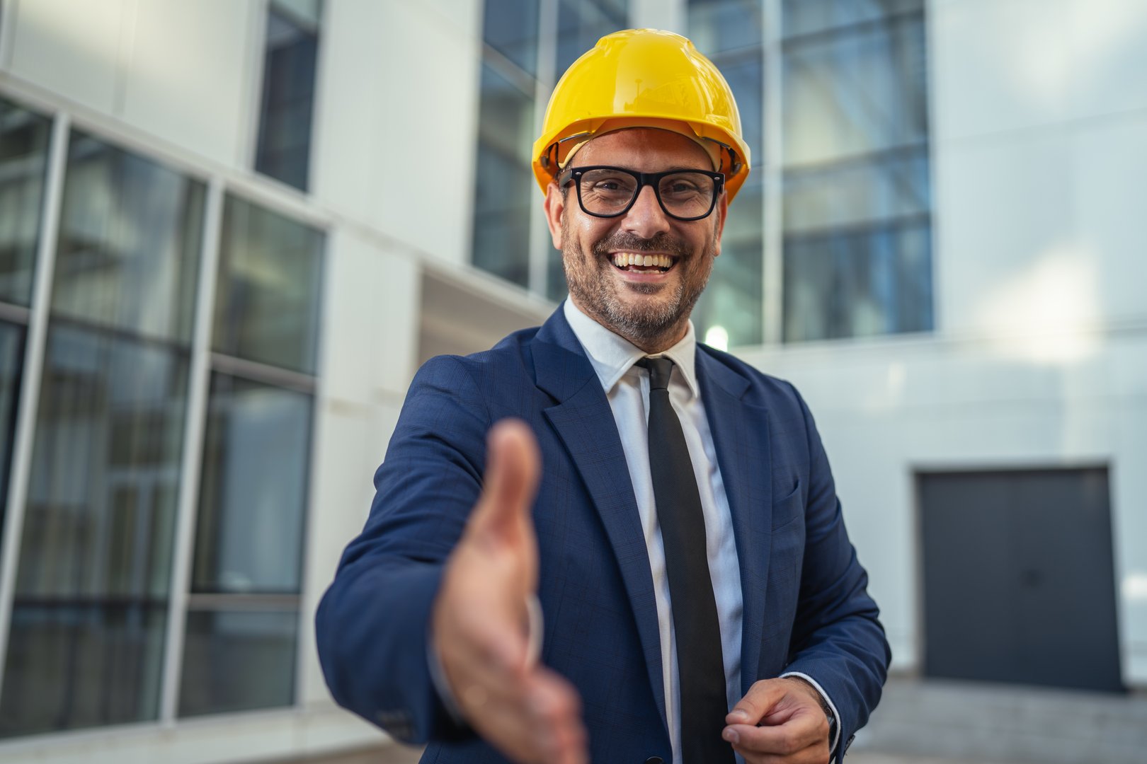 Confident male engineer or architect in a hard hat and suit extending an arm for a handshake, welcoming new partners or clients to a modern construction project