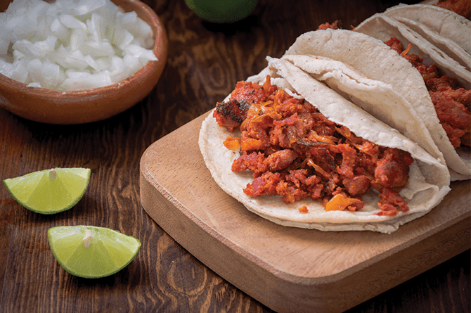 Two tacos with chorizo filling on a wooden board, with diced onions and lime wedges nearby.